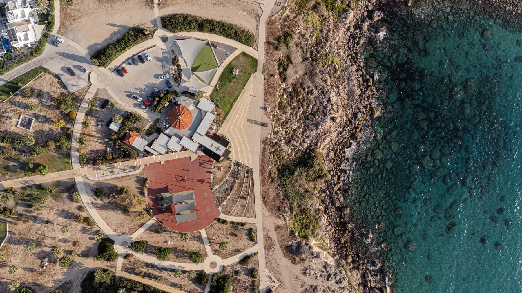 Top-down aerial of the promontory - the round red-tiled roof, the red plaza, coastal rocks and turquoise water.