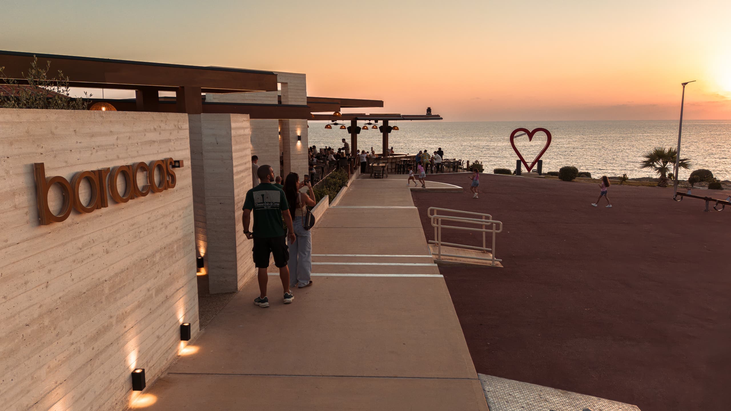 Guests walking in past the rust-coloured baracas wordmark on the travertine wall at sunset, the red heart sculpture and the plaza beyond.