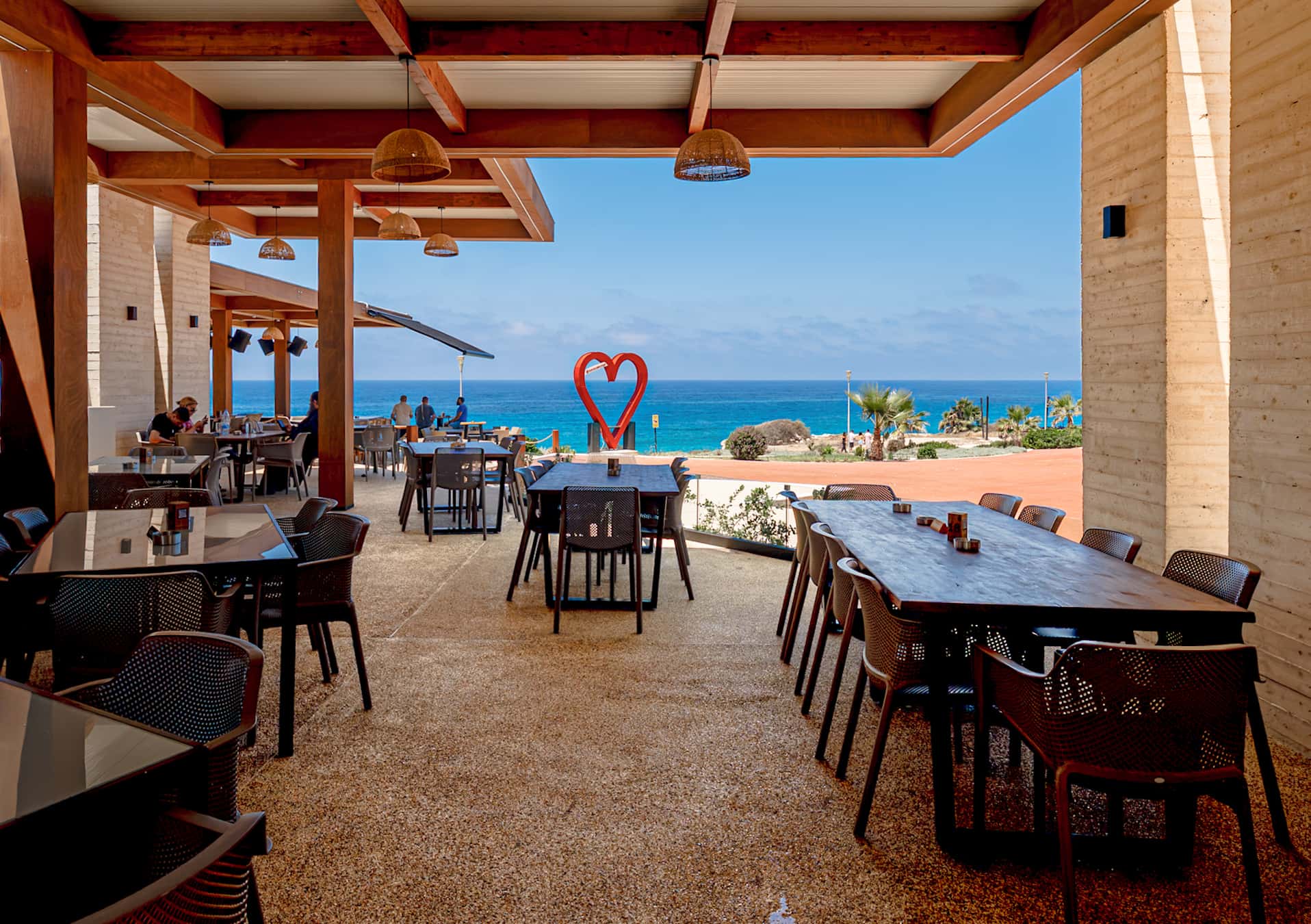 A long communal banquet table laid on the covered terrace, the red heart sculpture centred on the sea beyond.
