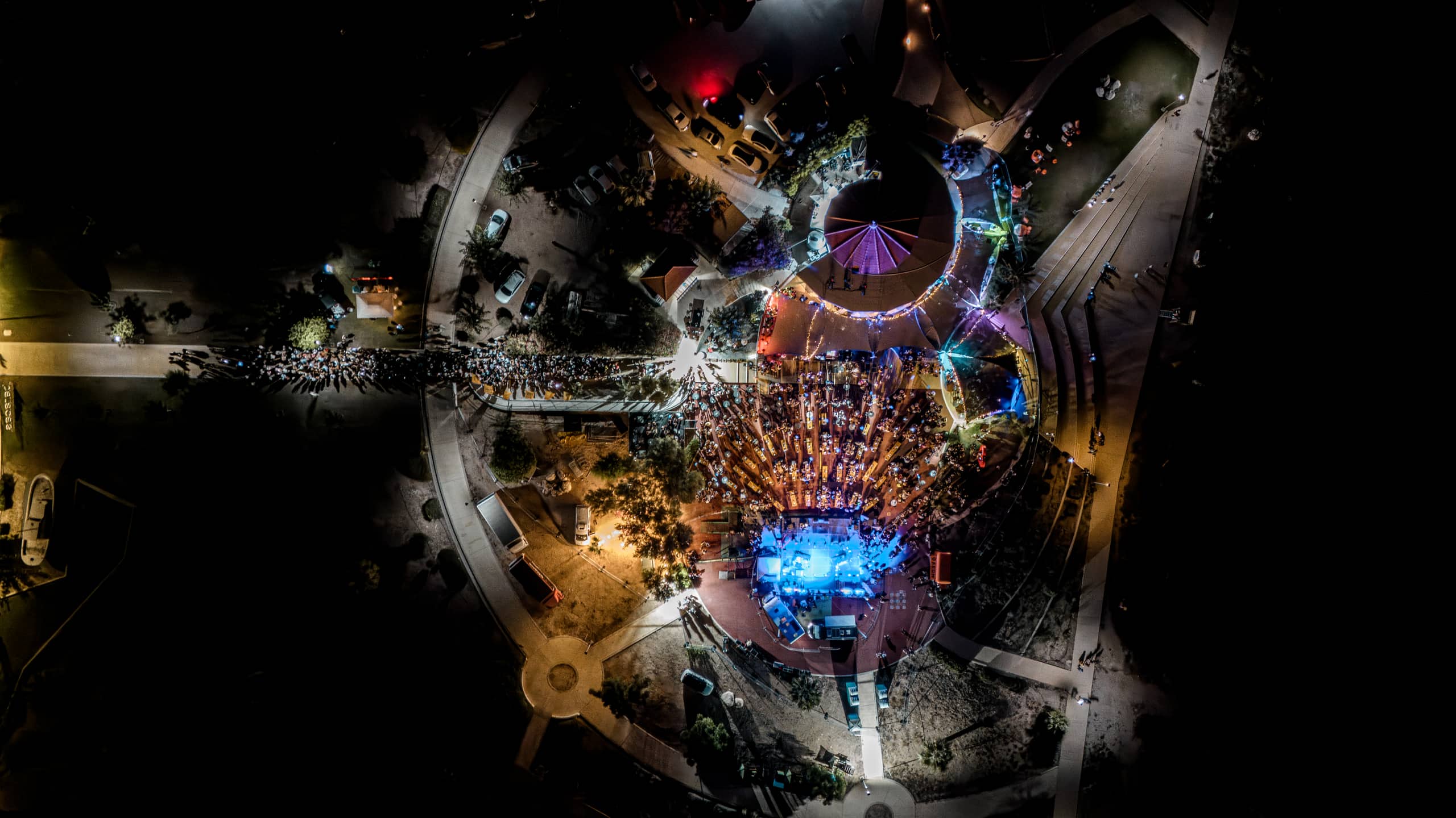 Aerial night view of a packed Baracas concert, the circular plaza and round roof lit in blue and coloured stage lights, a queue along the approach road and the dark coastline around it.