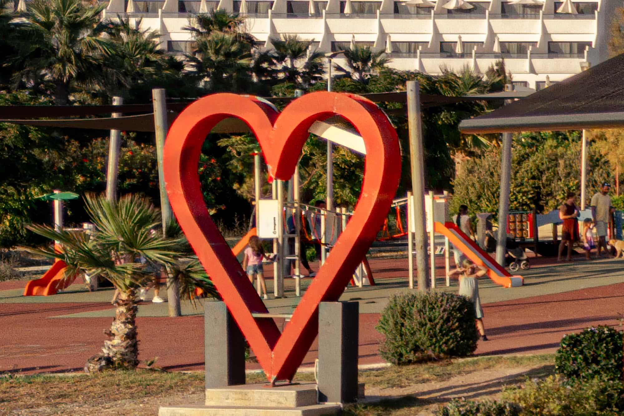 The red heart sculpture with the children's playground behind it - slides and climbing frames in use at golden hour.