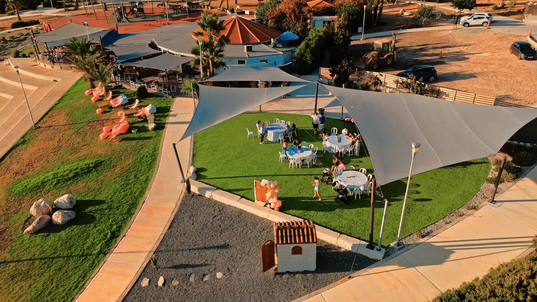 Aerial view of a private gathering on the Baracas lawn under white shade sails, round tables on the grass and the playground and round roof beyond at golden hour.