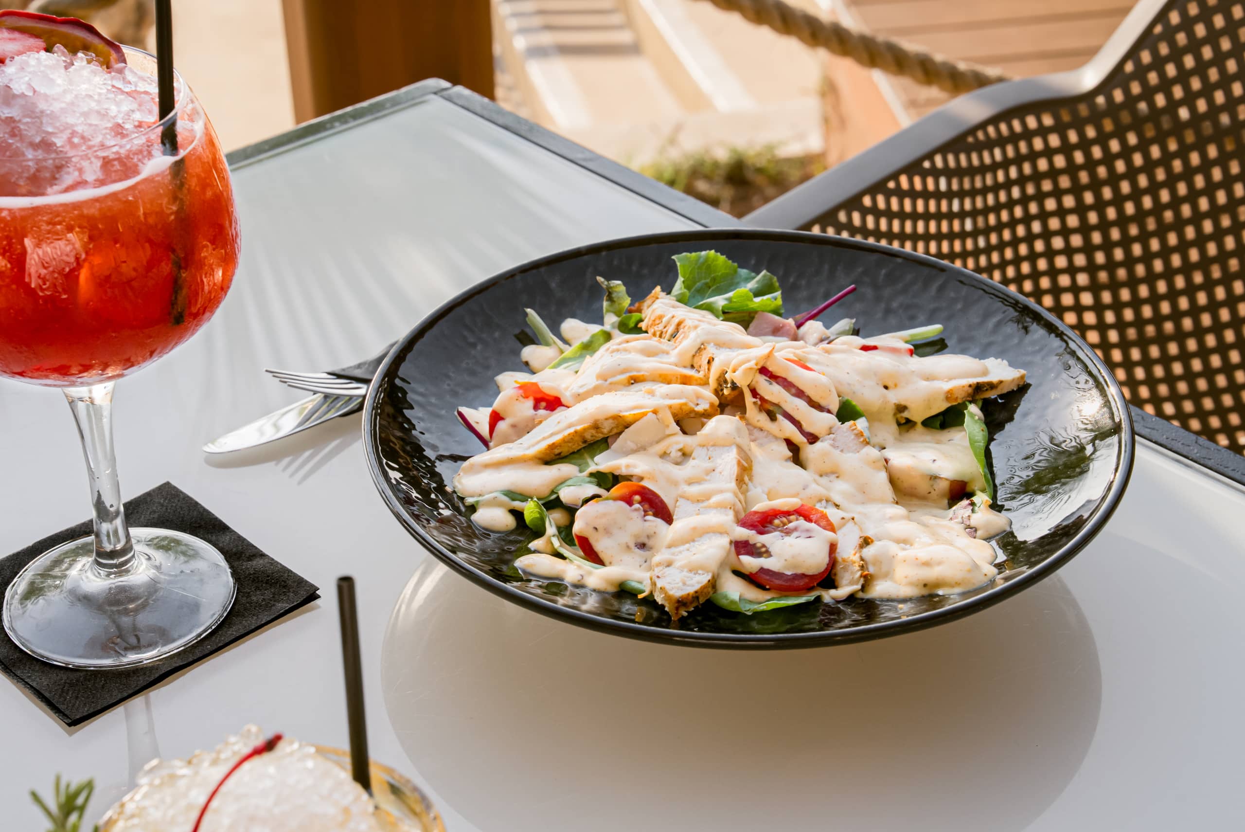 A grilled chicken salad in a black bowl beside a red spritz in a stemmed glass and a second cocktail on a terrace table.