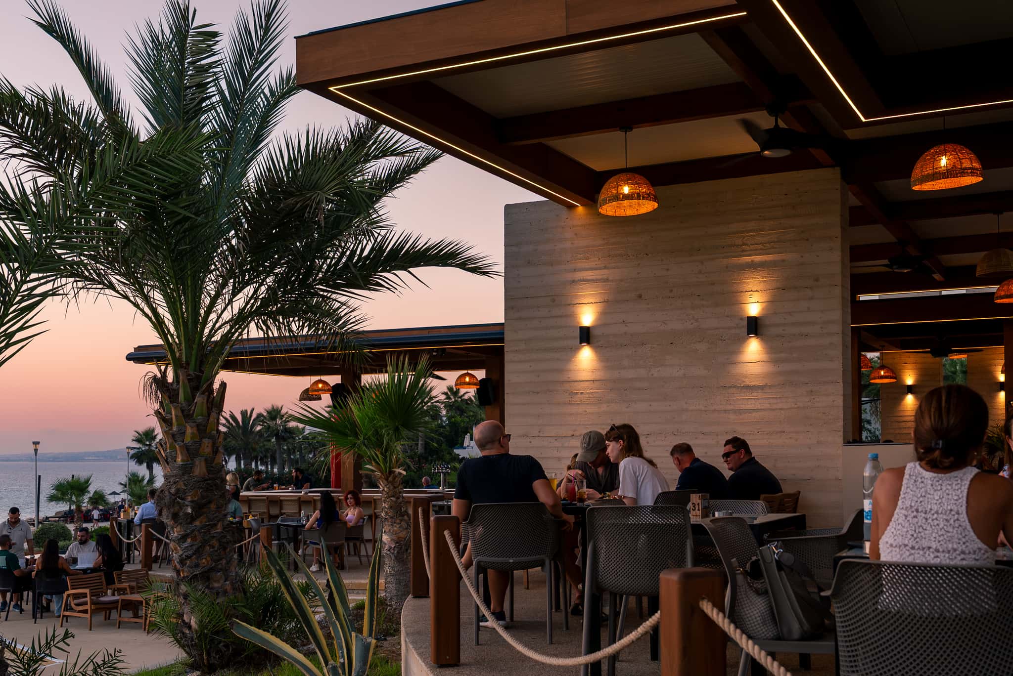 A dusk patio lit by two warm wall sconces on a raw concrete wall, guests seated at tables with the sea visible beyond.