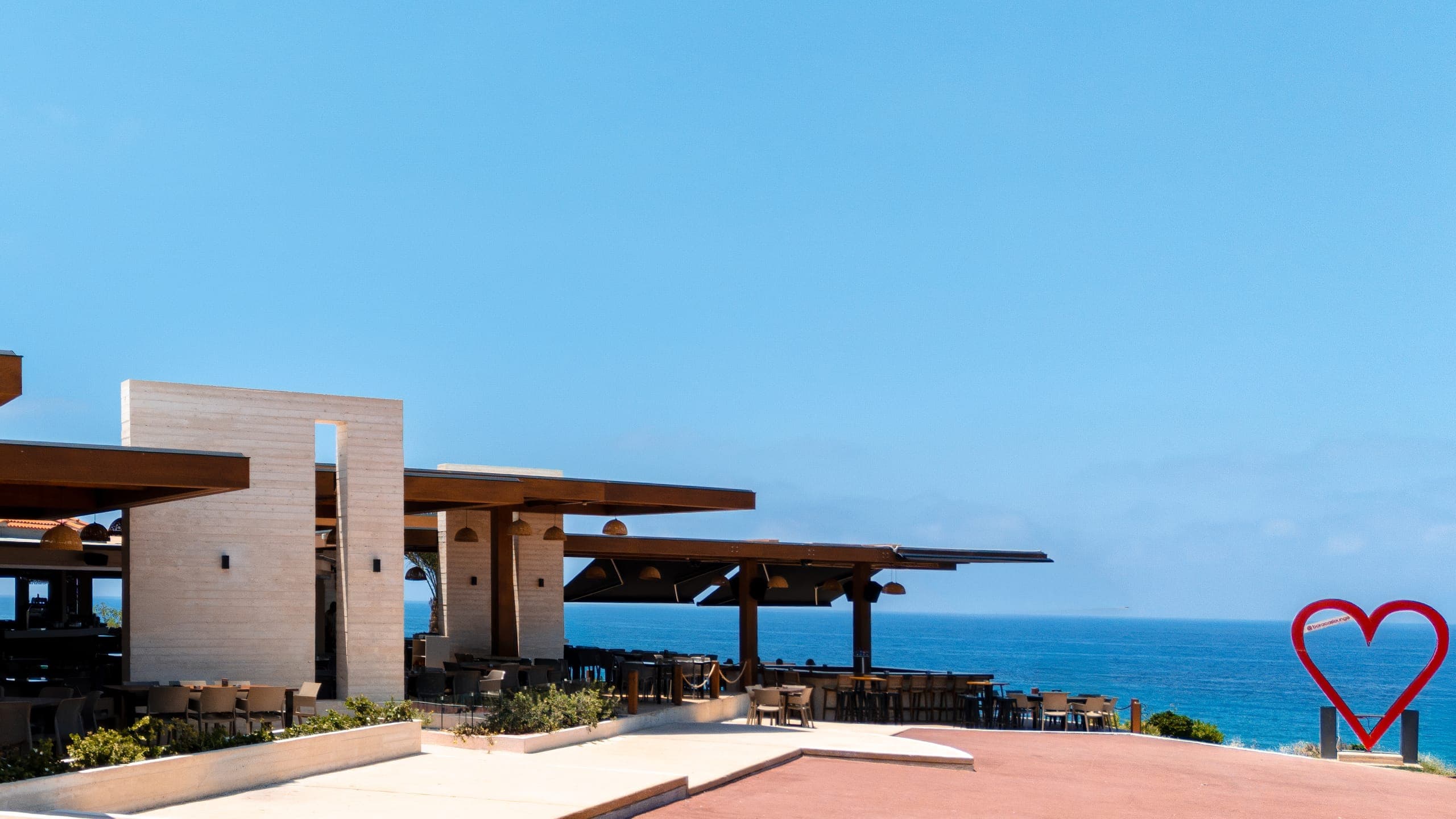 Wide daytime view of the Baracas building and pergola across the red plaza, the heart sculpture at the right under a deep blue sky.
