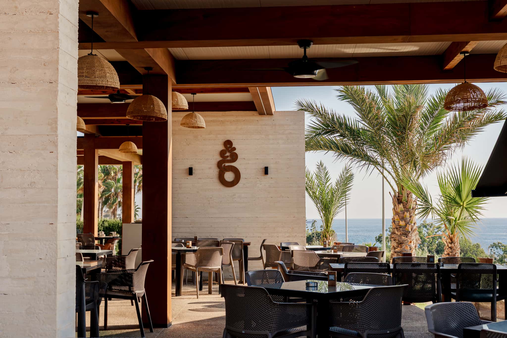 The covered daytime terrace - rattan pendant lamps, ceiling fans and the carved Baracas mark on the travertine wall, with palms and the sea beyond.