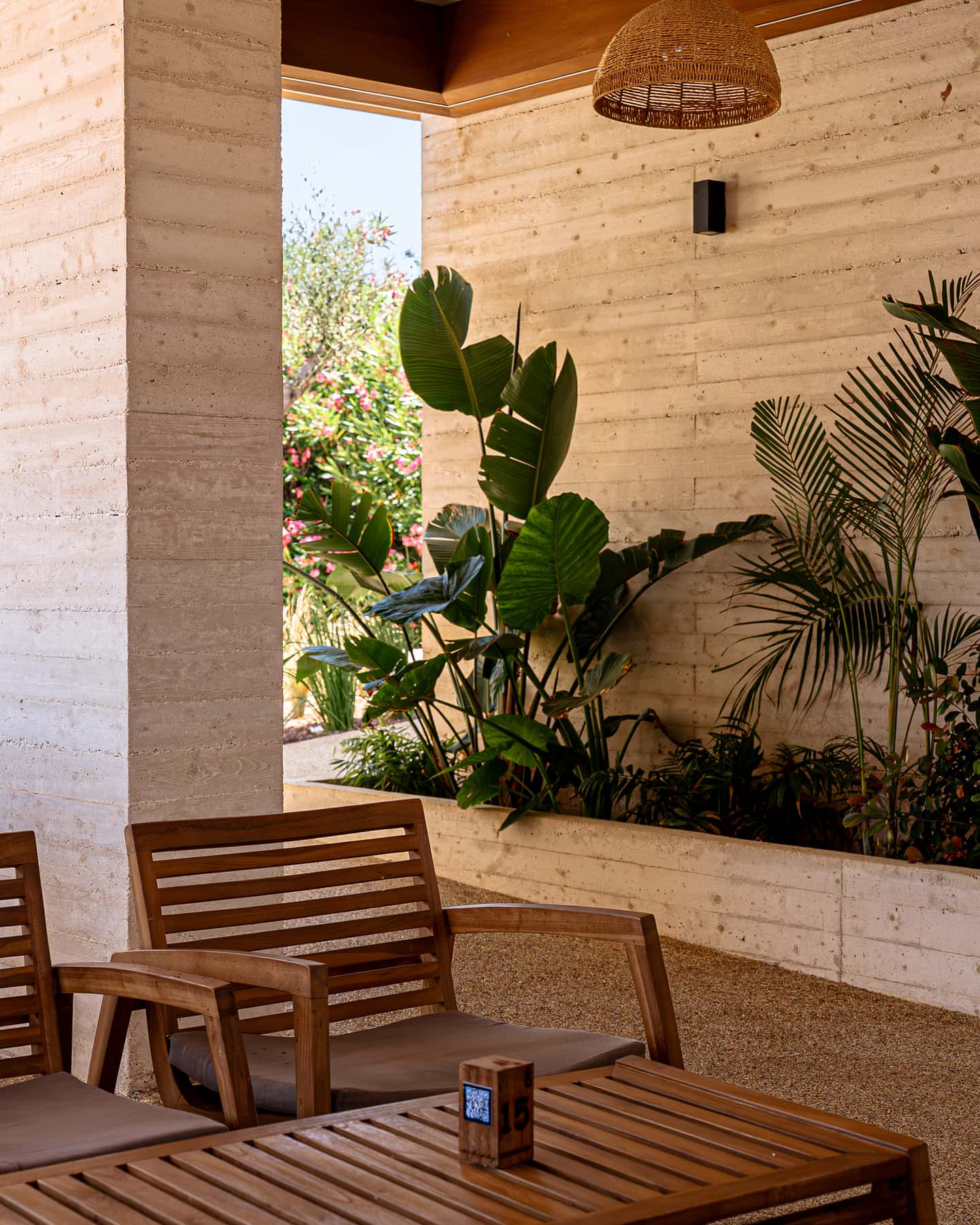 A shaded terrace with a teak bench, a bed of bird-of-paradise plants and a rattan pendant lamp overhead.