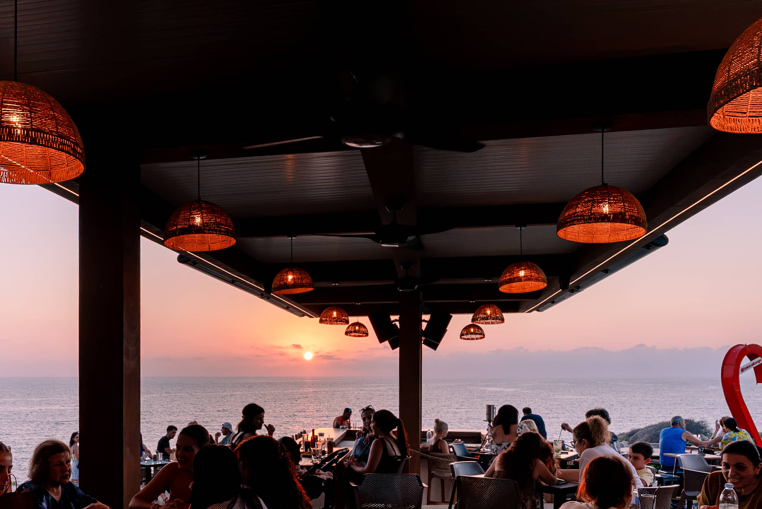 The packed Baracas terrace at sunset looking out to sea, rattan pendants glowing under the timber canopy and the red heart sculpture at the right edge.